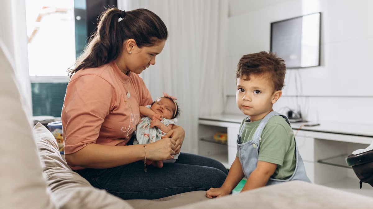 A mother sitting on a sofa with her baby and young son in a bright living room setting.